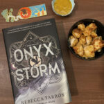 Physical hardcover version of ONYX STORM, photographed with a bookmark and a bowl of chicken nuggets as a snack